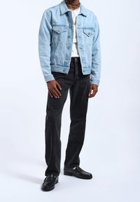 Chaqueta de denim azul claro con dos bolsillos frontales, llevada sobre una camiseta blanca, combinada con jeans negros y mocasines negros. Textura suave.