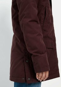 Burgunderfarbene Außenjacke mit glatter Textur, langen Ärmeln, seitlichen Taschen und Druckknopfverschluss am Saum.