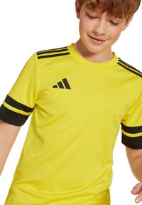 Gelbes Sportshirt mit schwarzen Akzenten, verfügt über schwarze Streifen an den Ärmeln und ein Logo. Hergestellt aus einem glatten, atmungsaktiven Stoff.