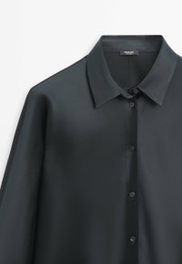 Chemise boutonnée en satin vert foncé avec col pointu, manches longues et étiquette de marque visible à l'intérieur du col.