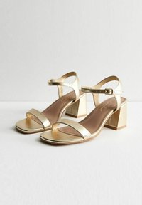 Sandalias de tacón bajo metálico dorado con dos tiras estrechas y una correa ajustable en el tobillo. Textura suave con un acabado brillante.