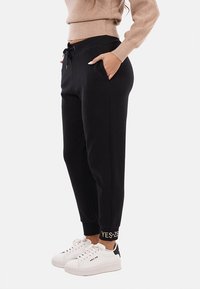 Pantaloni jogger neri con vita regolabile tramite cordino, tasche laterali e polsini a coste con il testo "YES". Il materiale è morbido e testurizzato. Abbinati a sneakers bianche.