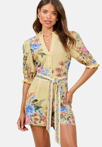 Floral mini dress in yellow with puff sleeves, featuring a button front, cinched waist, and a tie belt; decorated with various colorful flowers.