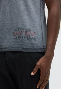Gray, short-sleeve t-shirt with a worn texture and red printed logo reading "Motorcycle Camp David Adventure" on the lower left.