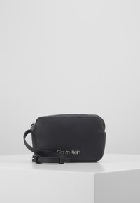 Černé kožené pouzdro obdélníkového tvaru, vybavené zipem a krátkým zápěstním poutkem. Přední strana zdobena stříbrným logem "Carly Klein".