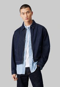 Giubbotto di jeans di colore blu scuro con design colletto, zip frontale e tasche laterali, indossato sopra una camicia azzurro chiaro con bottoni.