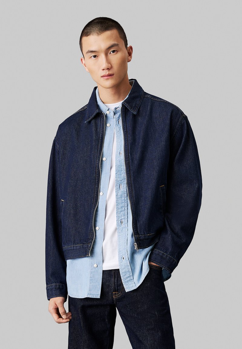 Giubbotto di jeans di colore blu scuro con design colletto, zip frontale e tasche laterali, indossato sopra una camicia azzurro chiaro con bottoni.