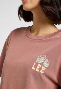 Rosa Baumwoll-T-Shirt mit kurzen Ärmeln, das ein Blumenmotiv und das Wort "LEE" in Beige auf der linken Brustseite präsentiert.