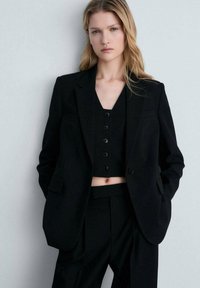 Černý elegantní blejzr s klopami, kombinovaný s odpovídajícím zkráceným topem na knoflíky a kalhotami s vysokým pasem, všechny v hladkém materiálu.
