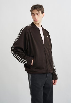 Jeune homme portant une veste noire zippée avec des bandes blanches sur les manches et un pantalon foncé à bandes latérales, regardant vers sa gauche.