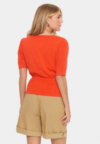 Vrouw die zijwaarts kijkt, gekleed in een oranje katoenen T-shirt met korte mouwen en geprinte mouwen, en beige kortere broekjes die opgerold zijn.