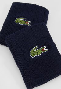 Duas pulseiras de pulso em tecido de felpa azul-marinho com bordado do logotipo de crocodilo verde, apresentando detalhes vermelhos e superfície texturizada.