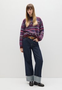 Burgunderfarbener Pullover mit mehrfarbigen Mustern, kombiniert mit dunklen, weiten Jeans mit umgeschlagenem Saum aus Denim. Brauner Gürtel und schwarze Schuhe.