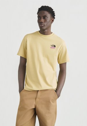 Young man with hands in pockets wearing a pale yellow The North Face 1966 logo t-shirt and light brown pants on a plain background.