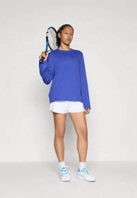 Γυναίκα τενίστρια στέκεται κρατώντας τη ρακέτα πάνω από τον ώμο, φορώντας μπλε μακρυμάνικο μπλουζάκι, άσπρα σορτς, άσπρες κάλτσες και άσπρα αθλητικά παπούτσια.