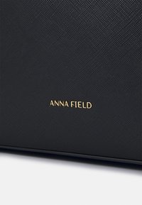 Black textured handbag featuring a gold "ANNA FIELD" logo. The surface has a subtle grid pattern with a structured shape.