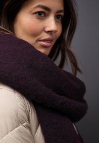 Une écharpe épaisse et violet foncé tombe autour d'une doudoune beige clair, mettant en valeur une texture douce et un design lâche en couches.