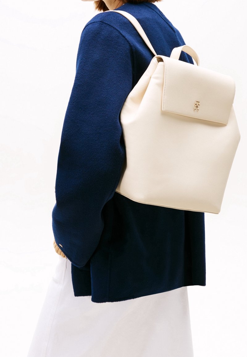 Personne portant un sac à dos en cuir crème sur un pull bleu foncé et une jupe ou un pantalon clair, vue de dos et de côté.