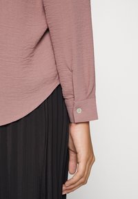 Blusa a maniche lunghe di un leggero color mauve con tessuto testurizzato e polsini abbottonati, abbinata a una gonna nera plissettata.