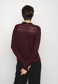 Maglietta borgogna a manica lunga con un design di pizzo a ricciolo sulla schiena e una texture liscia. Indossata con pantaloni neri, mani incrociate dietro.