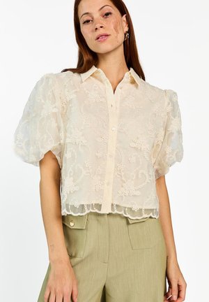 Femme portant un chemisier en dentelle crème à manches bouffantes et un pantalon taille haute kaki clair avec poches avant boutonnées.