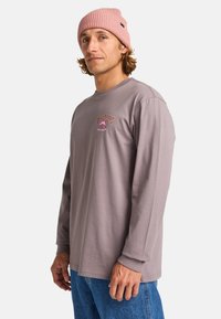 Szara koszula z długim rękawem z haftowanym różowym wzorem, w połączeniu z niebieskimi dżinsami i różowym, prążkowanym beanie, prezentująca luźny krój.