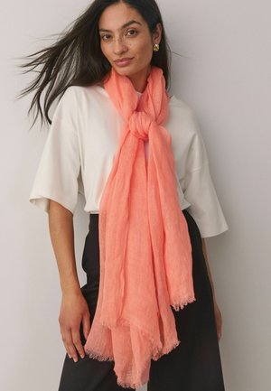 Woman with long dark hair wears white short-sleeve top, black pants, and a large coral scarf tied around her neck, posing against a plain wall.