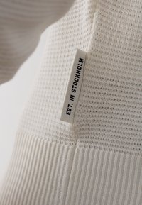 Weißer gestrickter Pullover mit strukturiertem Muster. Mit geripptem Saum und einem Etikett, auf dem "EST. IN STOCKHOLM." steht. Weiches Material.
