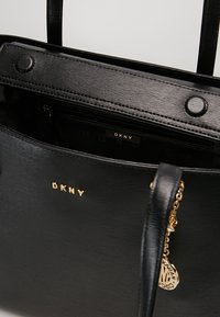 Czarna torba tote DKNY o teksturowanym wykończeniu, z złotym logo, dwoma górnymi uchwytami, wewnętrzną kieszenią na zamek oraz złotym akcentem w postaci zawieszki.