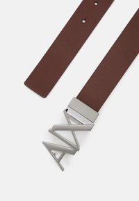 Brauner Leder-Gürtel mit strukturierter Oberfläche und einer silbernen Metallschnalle, die wie stilisierte Buchstaben geformt ist und ein elegantes rechteckiges Design aufweist.