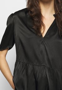 Blusa de satén negro con escote en V, mangas cortas y cintura fruncida. La tela tiene una textura suave y un brillo sutil.