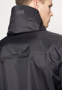 Chaqueta impermeable negra con un tejido texturizado, cuello alto y un logo en relieve en la parte trasera. Presenta costuras visibles y un diseño sin costuras.