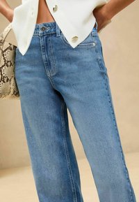 Világoskék farmernadrág, egyenes szabással, öt zsebes kivitelben, egyszerű gombos záródással. Az anyag sima tapintású.