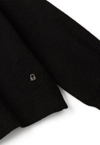 Schwarz gestrickter Pulloverärmel mit geripptem Bündchen und kleinem silbernen Logopatch, das in der Nähe des Bündchens auf einem weißen Hintergrund genäht ist.