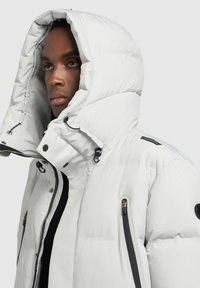 Fehér puffermellény nagy kapucnival, fekete cipzárakkal és kiegészítőkkel, steppelt textúrával és állítható zsinórral a galléren.