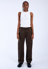 Witte geribbelde mouwloze top gecombineerd met bruin wijde broek. Model draagt zwarte schoenen. Eenvoudig en schoon ontwerp met neutrale kleuren.