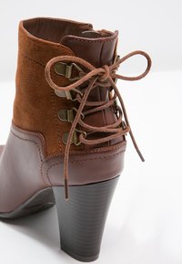 Bottine marron avec une tige en daim et des accents en cuir, présentant un design à lacets, des embellissements en métal et un talon haut empilé.