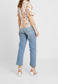 Blouse blanche avec un motif floral orange, manches courtes à volants ; jeans taille haute bleu clair avec ourlet effiloché ; sandales à talons carrés blanches.
