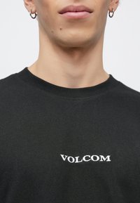 Κοντινό πλάνο προσώπου ενός ατόμου που φοράει μαύρο πουκάμισο με το κέντημα «VOLCOM» σε λευκό στο στήθος· το άτομο φοράει κρίκους στα αυτιά.