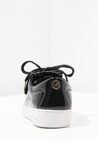 Sneaker in pelle verniciata nera con suola bianca, dotata di lacci neri, accentuazione metallica rotonda sul tallone e dettagli texture sulla parte superiore.