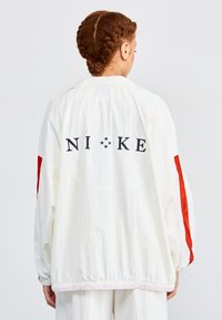 Fehér, könnyű dzseki piros részletekkel, hátul fekete "NIKE" logóval, rugalmas mandzsettákkal és összehúzható derékkal rendelkezik.