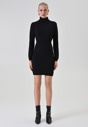 Donna che indossa un vestito aderente nero a collo alto e stivaletti neri con borchie, in piedi davanti a uno sfondo chiaro e semplice.