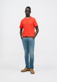 Rdeča bombažna majica s krožnim izrezom, kombinirana s svetlo modrimi kavbojkami. Obleko dopolnjujejo rjavi semiš čevlji z gumijastimi podplati.