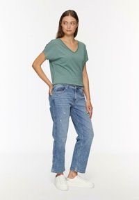 Groene v-hals T-shirt met korte mouwen, gecombineerd met lichtblauwe versleten jeans en witte sneakers. De outfit heeft een ontspannen pasvorm.