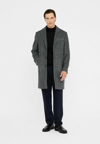 Cappotto di lana grey a spina di pesce con colletto a lancia, chiusura con bottoni e tasche laterali, abbinato a un dolcevita nero e pantaloni blu navy.