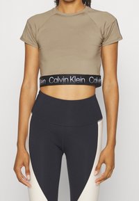 Brązowy krótki t-shirt z krótkimi rękawami i czarną gumką w pasie z logo Calvin Klein, zestawiony z czarno-kremowymi legginsami z wysokim stanem.