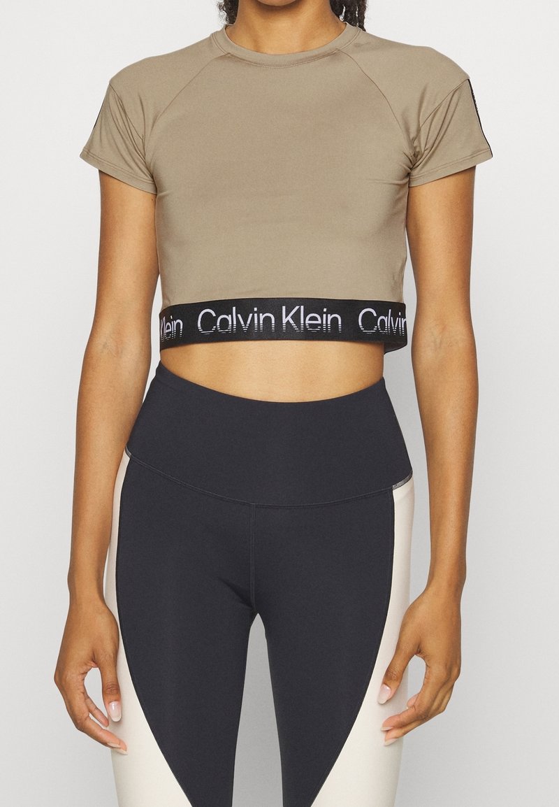 Brązowy krótki t-shirt z krótkimi rękawami i czarną gumką w pasie z logo Calvin Klein, zestawiony z czarno-kremowymi legginsami z wysokim stanem.