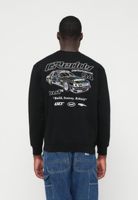 Mann trägt einen schwarzen Sweatshirt mit weißem Rennwagen-Graphic, Text "GReddy Performance 94," "Build, Destroy, Rebuild," und Logos auf dem Rücken, kombiniert mit blauen Jeans.