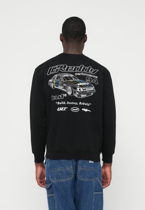 Mees kannab musta kapuutsiga särki, millel on valge võidusõiduauto graafika, tekst "GReddy performance 94," "Build, Destroy, Rebuild," ja logod seljal, koos siniste teksadega.