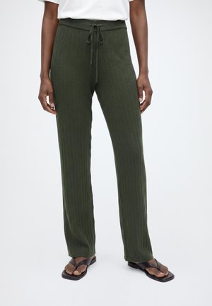 Personne portant un pantalon côtelé vert foncé avec un cordon et des sandales noires à bout carré, debout sur un fond blanc uni.